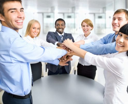 Eine gemischte Gruppe von sechs Personen steht in einem hellen Büro um einen Tisch herum, lächelt und legt in der Mitte ihre Hände aufeinander, was Teamwork und Zusammenarbeit symbolisiert. Sie tragen alle Business-Kleidung. Eine gemischte Gruppe von sechs Personen steht in einem hellen Büro um einen Tisch herum, lächelt und legt in der Mitte ihre Hände aufeinander, was Teamwork und Zusammenarbeit symbolisiert. Sie tragen alle Business-Kleidung.